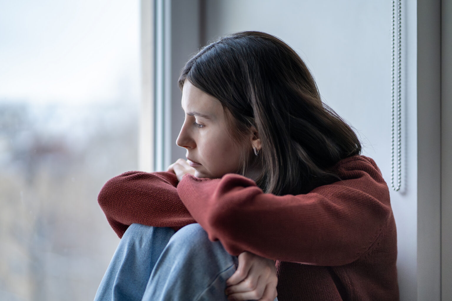 A young woman sitting at a window with her arms wrapped around her knees, looking out