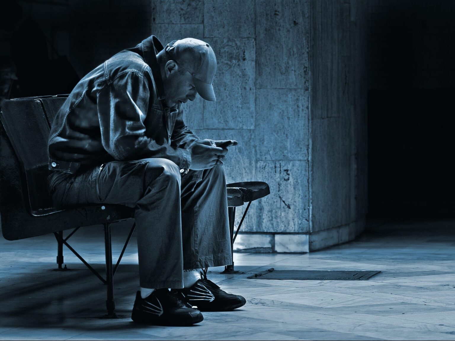 A person hunched over a phone on a bench in a dim, empty underpass